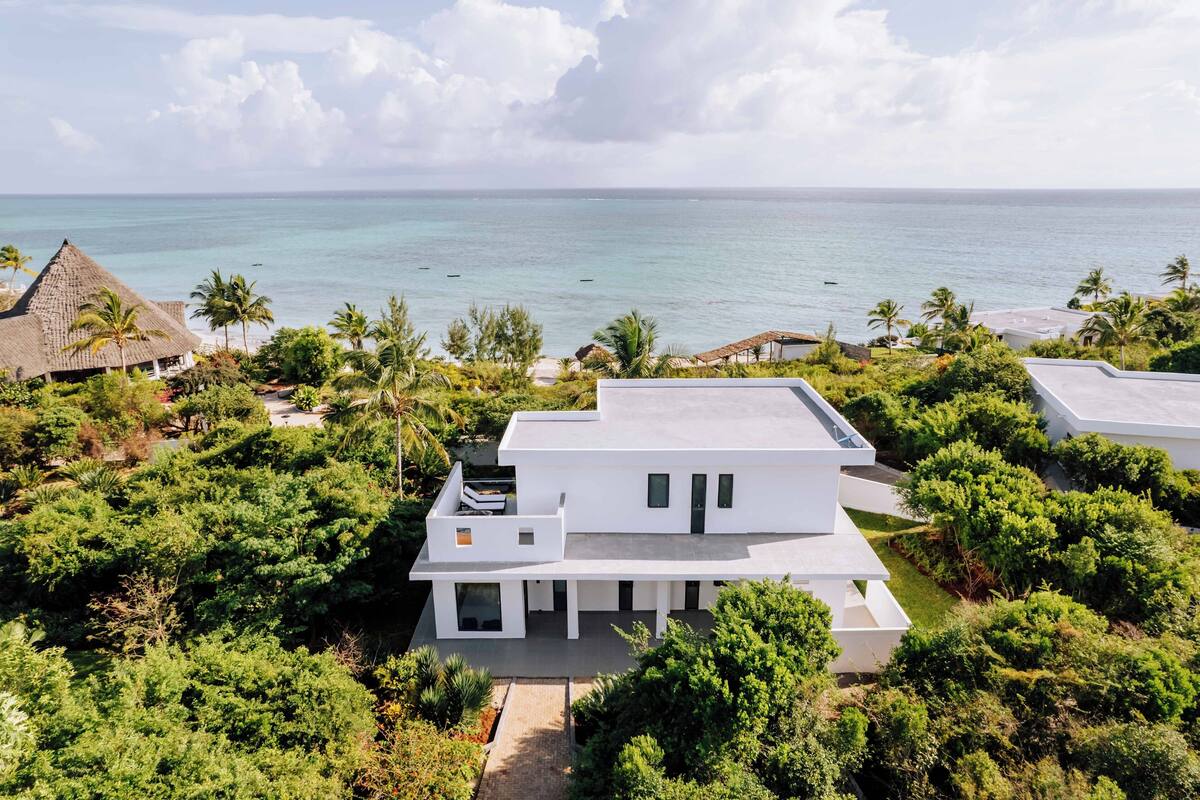 A modern white building with a flat roof, surrounded by lush greenery and overlooking a calm sea.