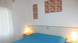 Une chambre avec un lit simple, deux tables de chevet avec des lampes, une fenêtre avec un rideau et deux éléments de décoration florale sur les murs.