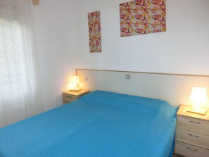 Quarto com cama, duas mesas de cabeceira com luminárias, parede com duas imagens de padrão floral e janela com cortina branca.