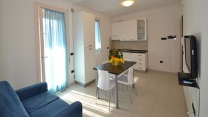 A modern living room with a blue sofa, a dining table with chairs, a kitchen area with white cabinets, and a TV mounted on the wall. - Modern Apartment by the Beach (Rosolina)
