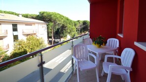 A balcony with white plastic chairs and a table, red walls, and a view of buildings and trees. - Modern Apartment by the Beach (Rosolina)