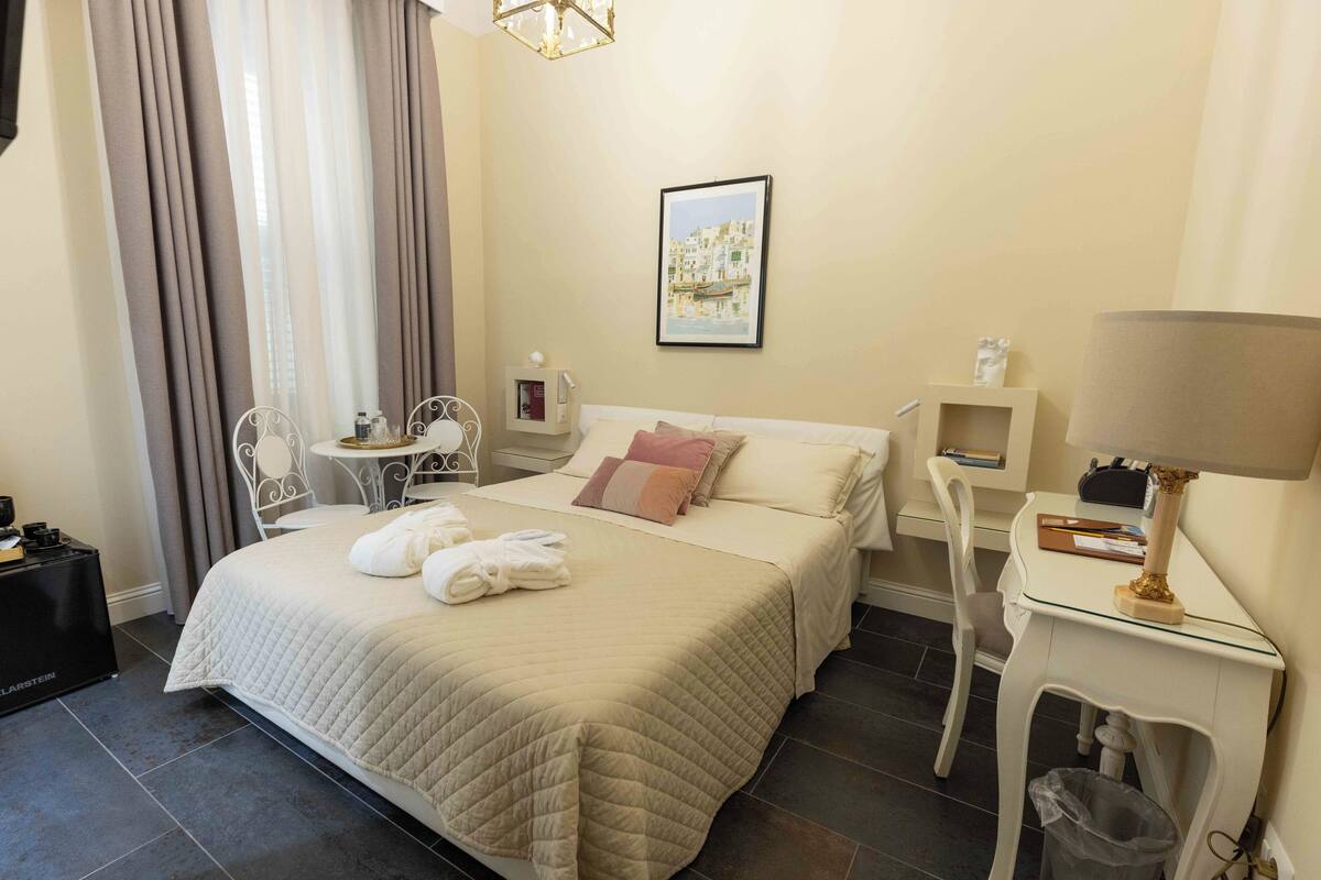 A hotel room with a bed, bedside table, chair, and a desk with a lamp.
