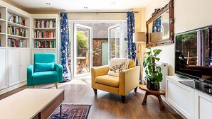 A living room with a bookshelf, a teal armchair, a yellow armchair, a television, and a large window with curtains. - Terraced Bermondsey House (London)