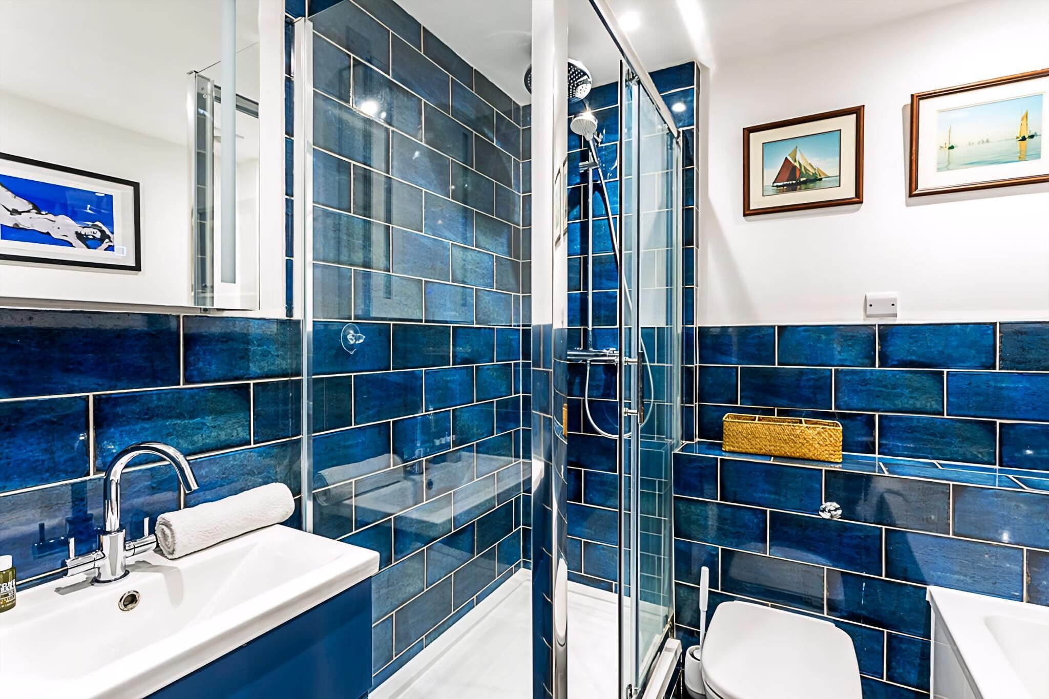 A bathroom with blue-tiled walls, a glass-enclosed shower, a white sink, and two framed pictures on the wall.