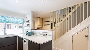 A modern kitchen with dark cabinetry, a white countertop, and a view of a staircase leading to a landing. - Terraced Bermondsey House (London)