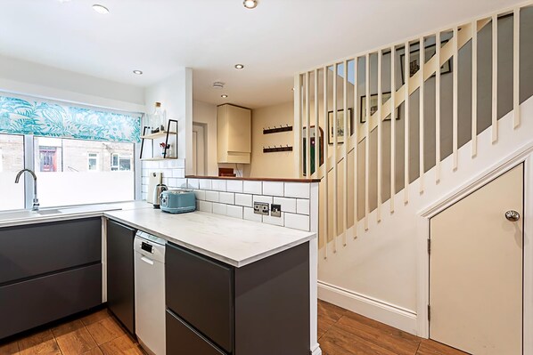 A modern kitchen with dark cabinetry, a white countertop, and a view of a staircase leading to a landing.