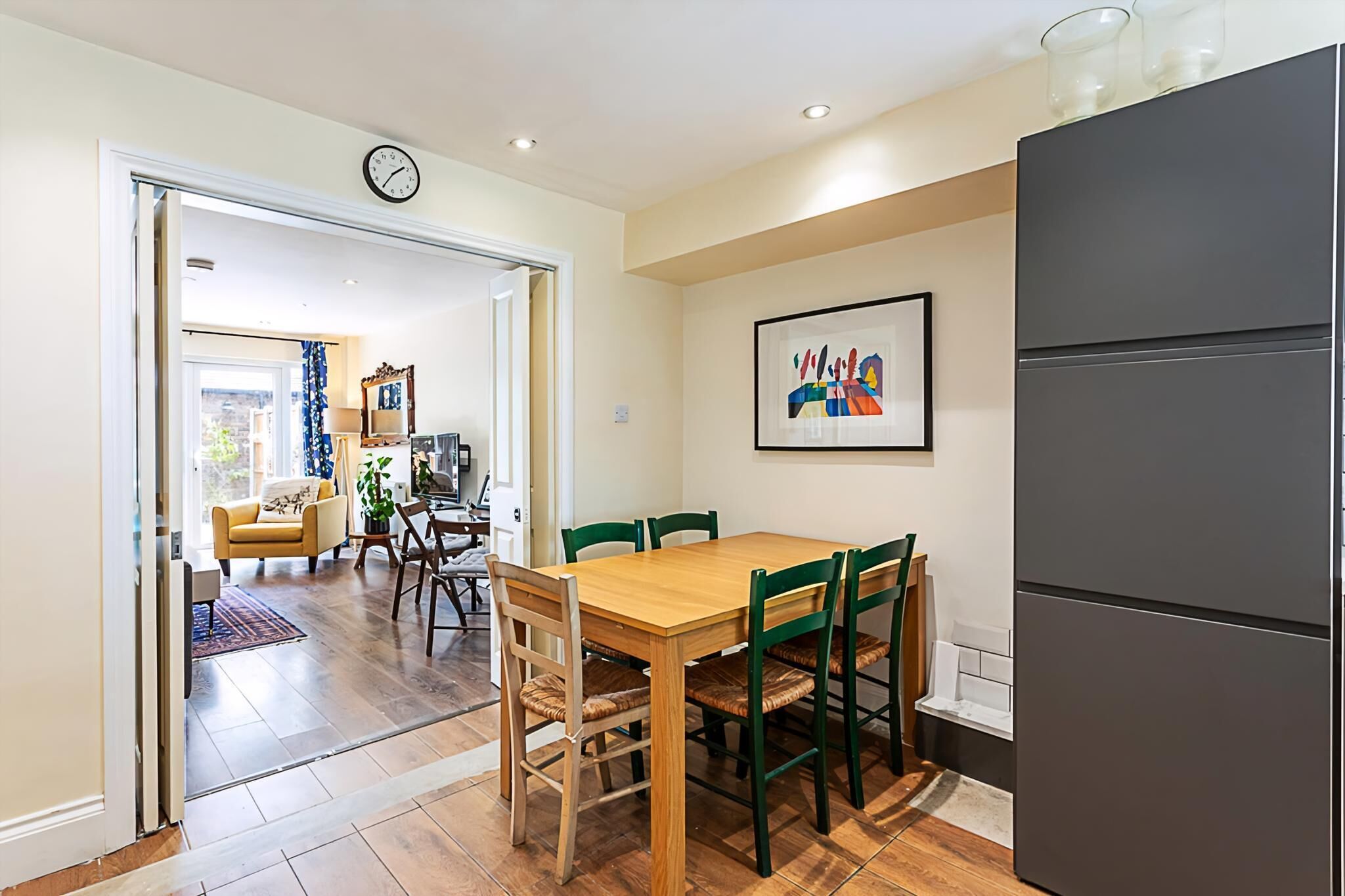 A modern kitchen with a wooden table and green chairs, a grey refrigerator, and a clock on the wall.