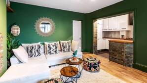 Una sala de estar moderna con una pared verde de acento, un sofá blanco y una cocina visible a través de un vano abierto.