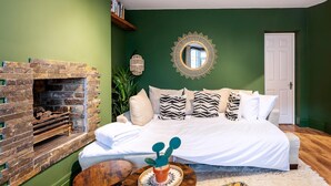 Un dormitorio con chimenea de ladrillo, una cama con ropa blanca, almohadones estampados, un espejo redondo y una puerta.