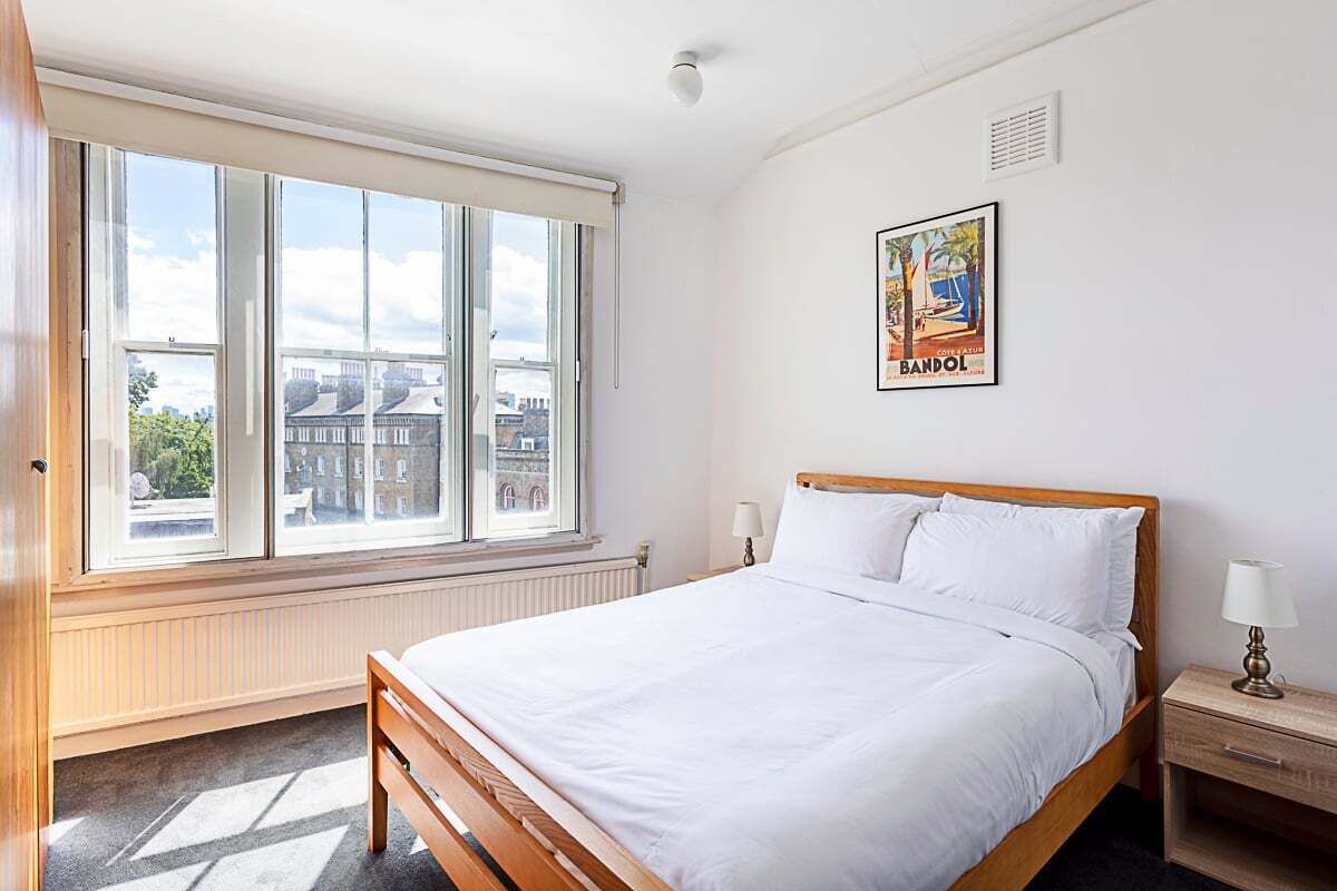 A bedroom with a large window, a bed, a bedside table, and a wall-mounted lamp.