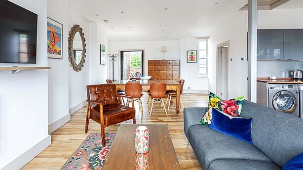 A spacious living room with a grey sofa, wooden coffee table, and a dining area with a table and chairs. - Contemporary Open Plan Penthouse (London)