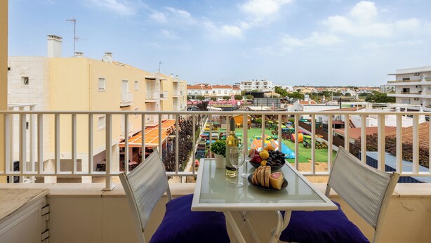 Un balcón con una mesa dispuesta para dos personas, con vistas a una zona residencial con edificios de colores y un cielo despejado.