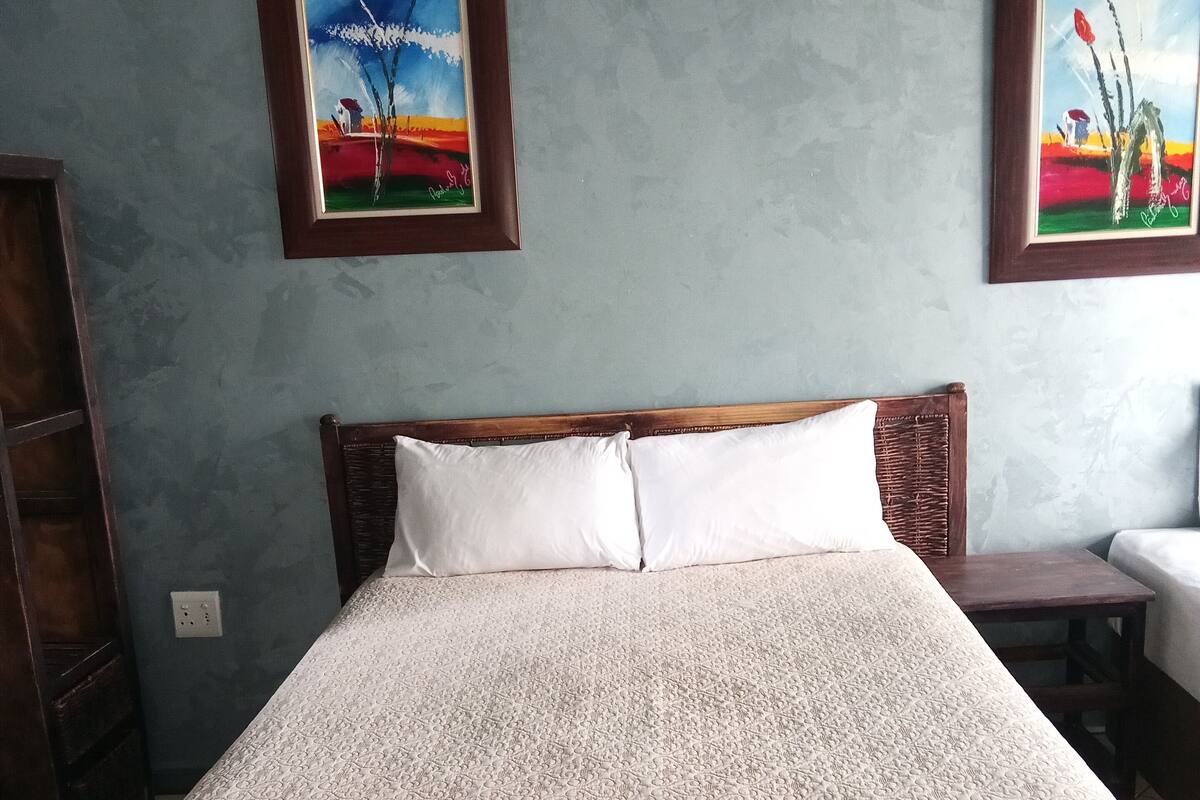 Een bed met een bedsprei met dessin, een houten hoofdbord en twee schilderijen aan de muur.