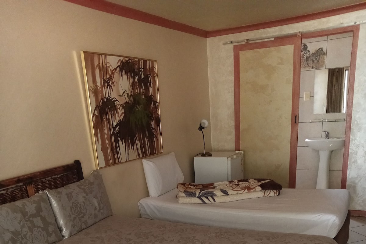 Een slaapkamer met twee bedden, een nachtkastje, een lamp, een schilderij en een badkamer met een wastafel en spiegel.