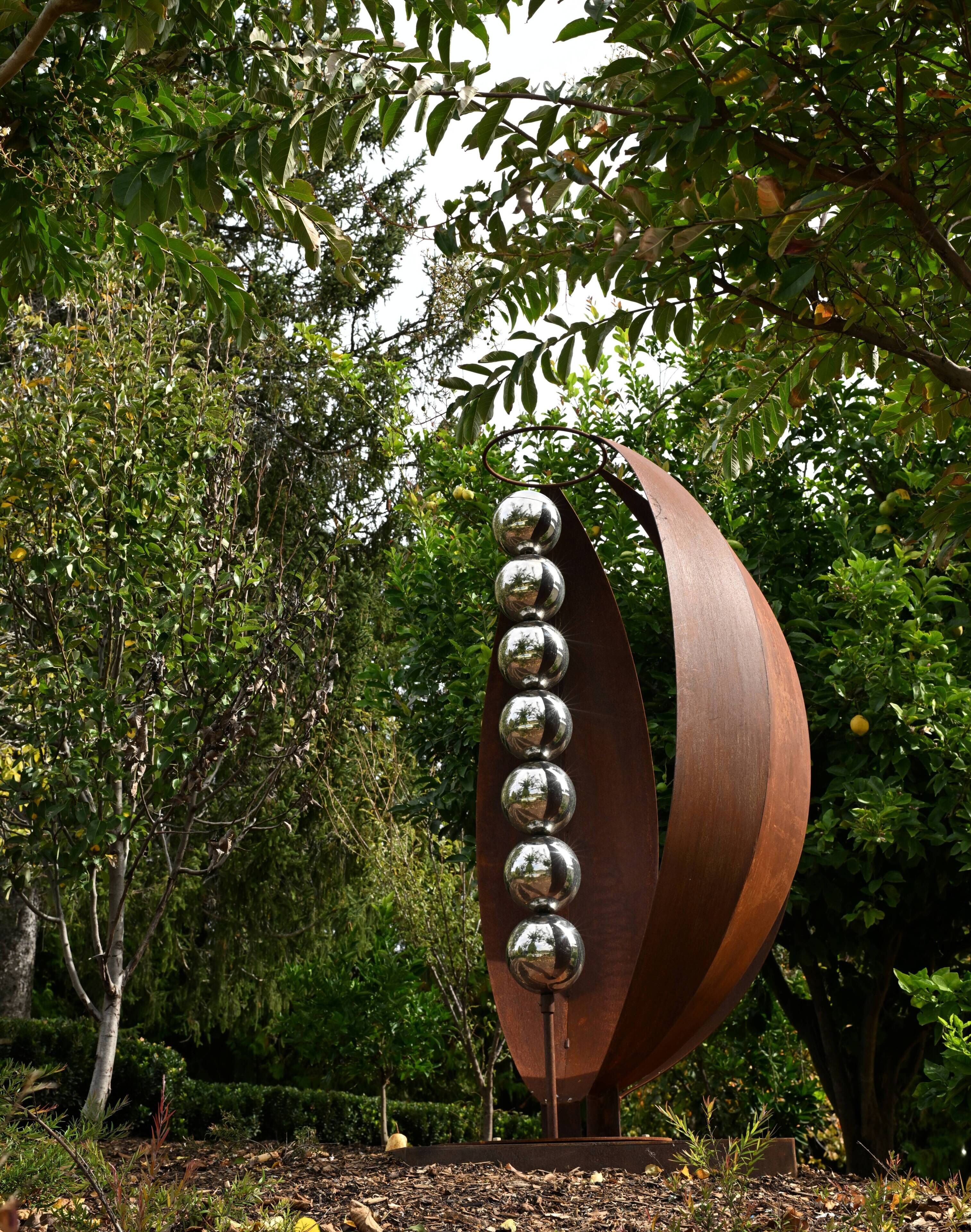 A metal sculpture with a series of reflective spheres mounted on a curved structure, set in a garden with trees and shrubs.