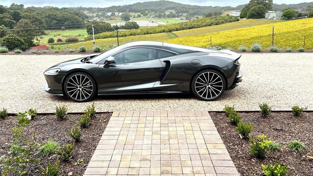 A sleek, dark-colored sports car parked on a gravel driveway with a brick pathway leading to it.