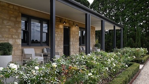 Un bâtiment en pierre doté d’un patio couvert, d’un espace extérieur aménagé pour s’asseoir et d’un jardin orné de fleurs blanches.