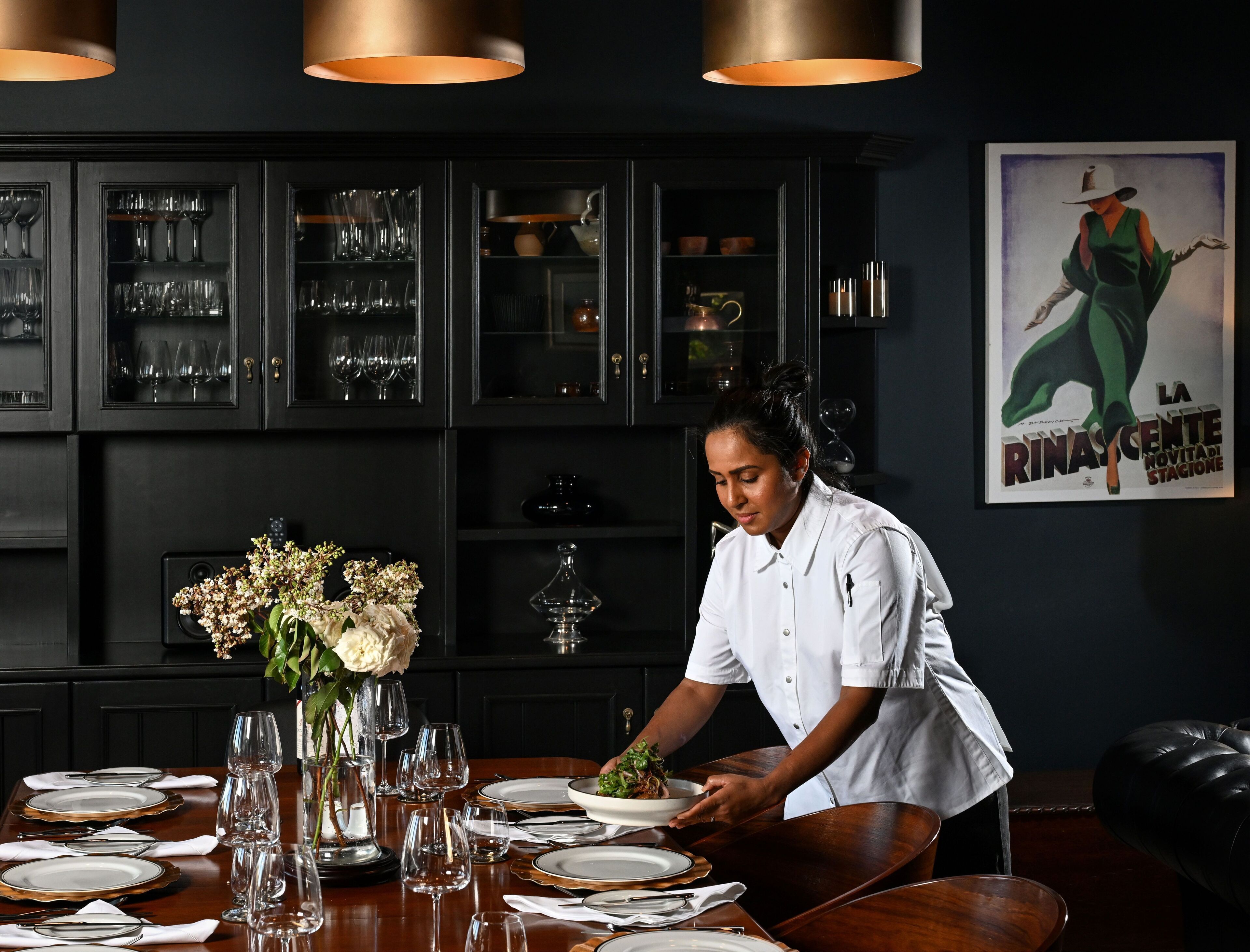 Un espace repas où un chef prépare les plats, une table dressée avec des assiettes et des verres, et un intérieur sombre agrémenté d’armoires aux façades en verre.