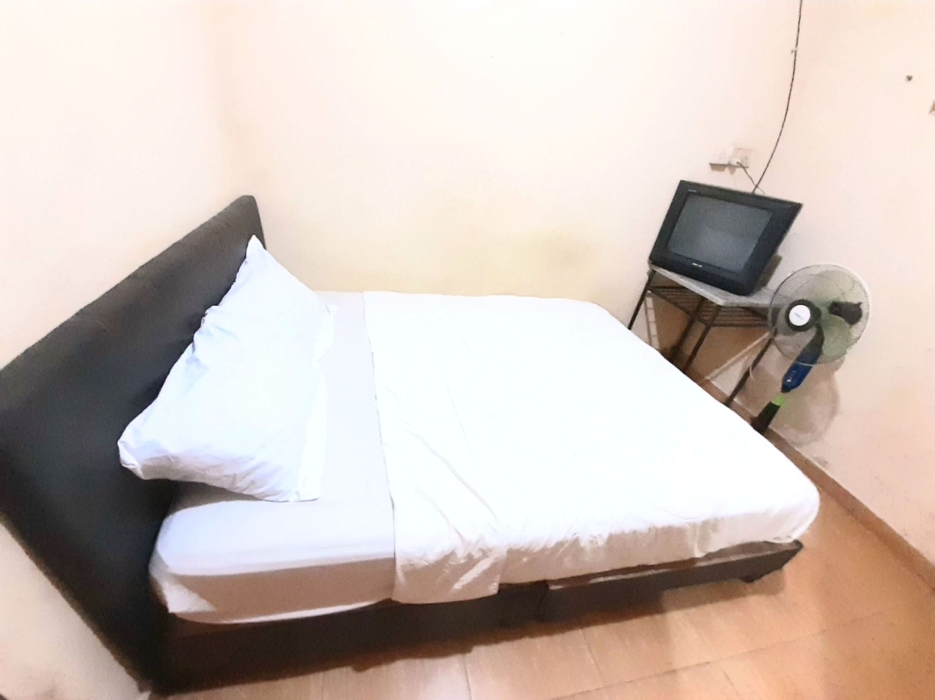 Ein Einzelbett mit weißer Bettwäsche, ein schwarzer Kopfteil, ein kleiner Tisch mit Fernseher und Ventilator sowie eine schlichte Wand mit Steckdose.