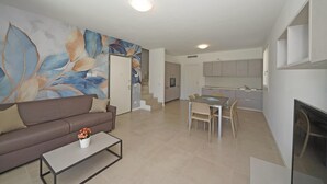 Una sala de estar moderna con un sofá, una mesa de centro y un cuadro en la pared. Se observa un comedor con una mesa y sillas, y una cocina al fondo.