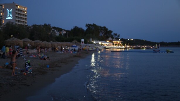 A beach with umbrellas and people, a hotel with a blue logo, and a body of water reflecting lights.