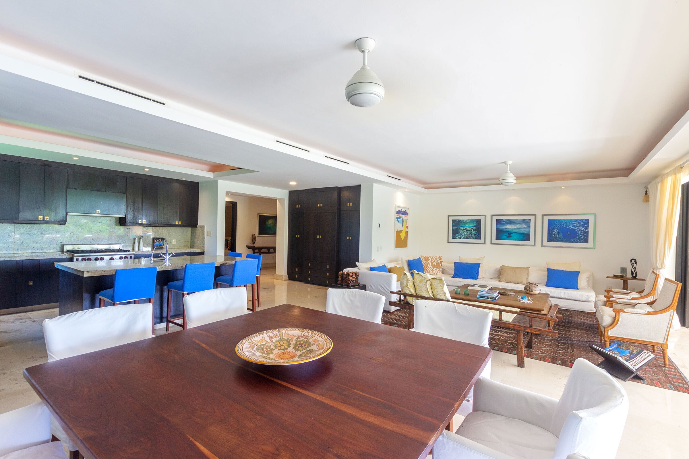 Uma cozinha moderna com armários escuros, uma ilha central e cadeiras azuis. Há uma mesa de jantar com uma tigela decorativa no centro. A sala de estar apresenta um conjunto de sofás, uma mesa de centro e obras de arte emolduradas na parede.