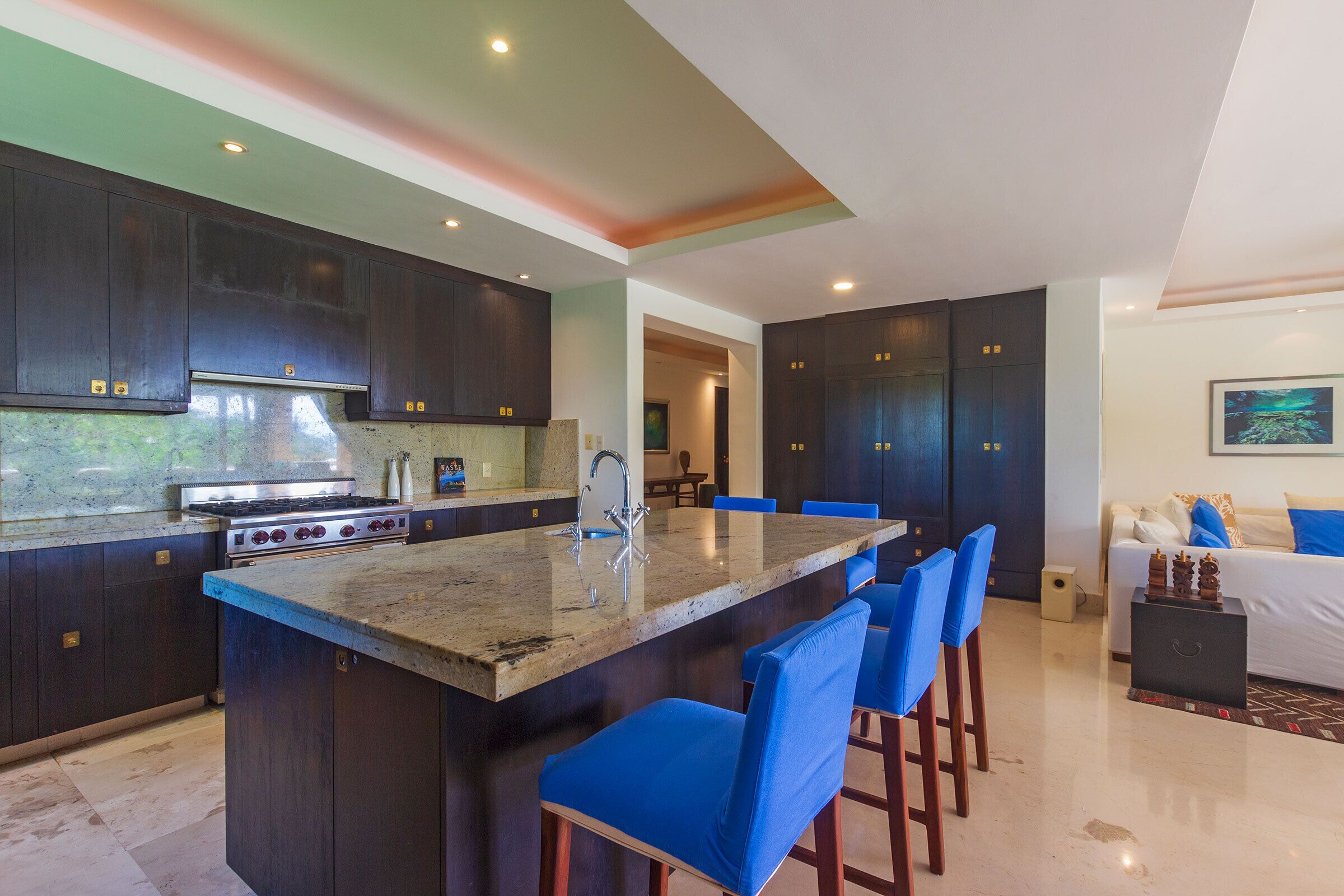 Een moderne keuken met donkere kastjes, een centraal eiland met blauwe stoelen, en een eethoek met een bank en een schilderij aan de muur.