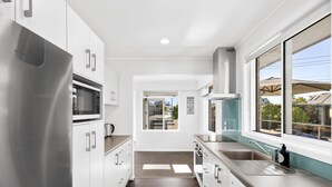 A modern kitchen with stainless steel appliances, white cabinetry, and a view of the neighborhood through large windows. - Mount Maunganui Beach House Family Friendly (Mount Maunganui)
