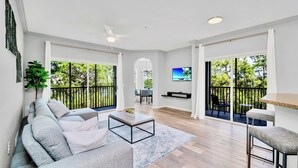 A modern living room with a sofa, coffee table, and TV. There are large windows with a view of trees and a balcony. - Upstay Pet-Friendly Condo Near Disney w Balcony (Winter Garden)