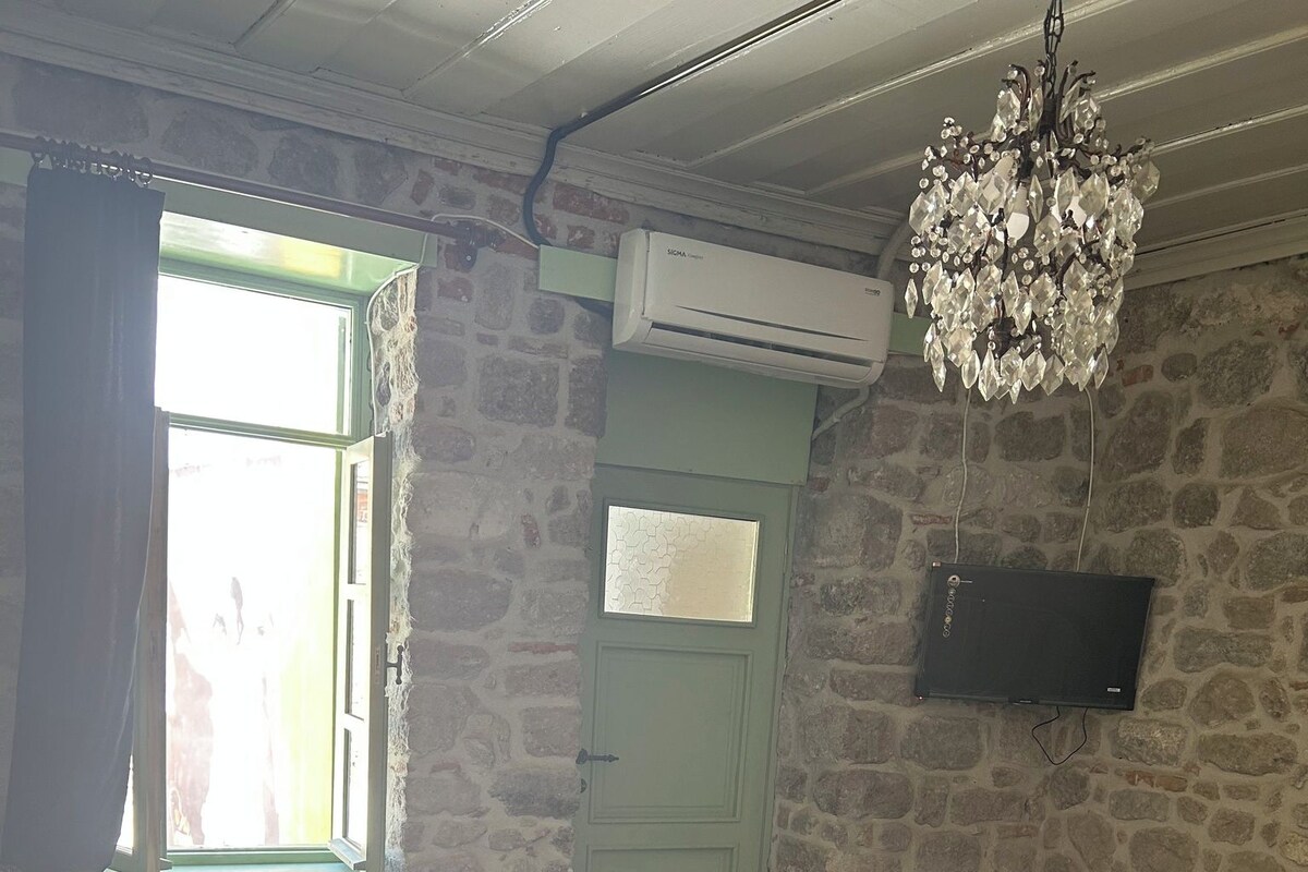 Een kamer met een stenen muur, een bed met een groene sprei, een witte stoel, een rood tapijt en een kroonluchter.