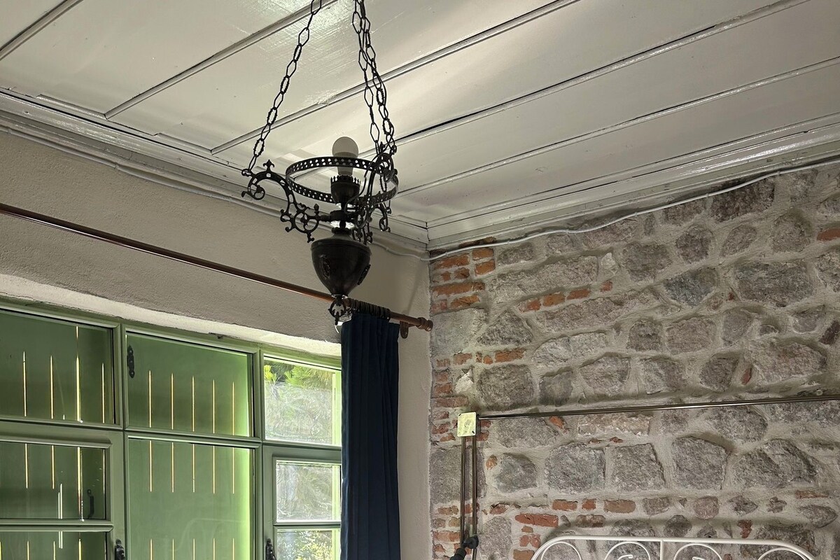 Une chambre avec un mur en pierre, un cadre de lit en métal, une fenêtre verte et un luminaire suspendu.