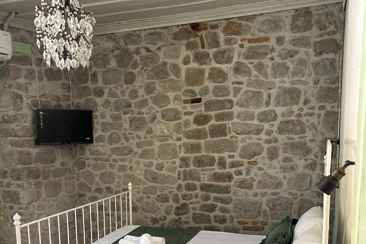 Una cama individual con estructura blanca, una manta verde y un almohadón blanco. Se aprecia una pared de piedra detrás de la cama y una lámpara de araña colgada arriba.