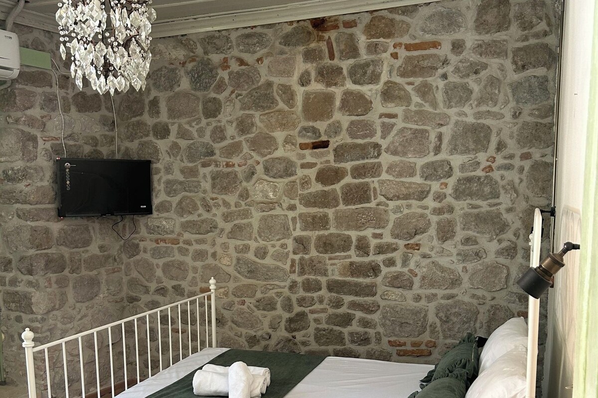 Un lit avec un cadre blanc, une couvre-lit verte et un plaid vert foncé. Un lustre est suspendu au plafond, et un téléviseur à écran plat est fixé au mur en pierre.