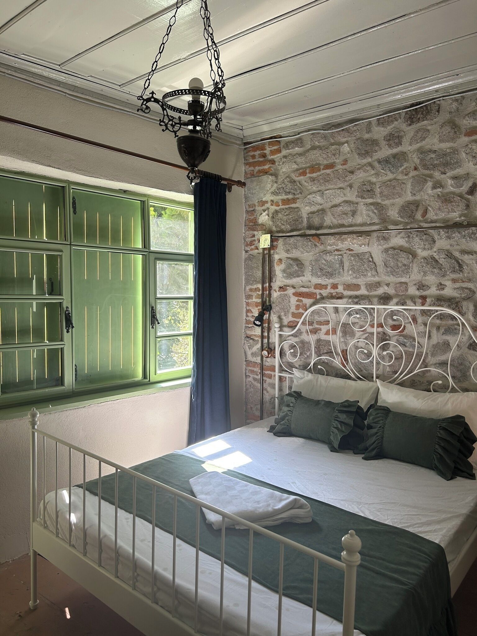 Een slaapkamer met een stenen muur, een metalen bed, een groen raam en een hangende lamp.