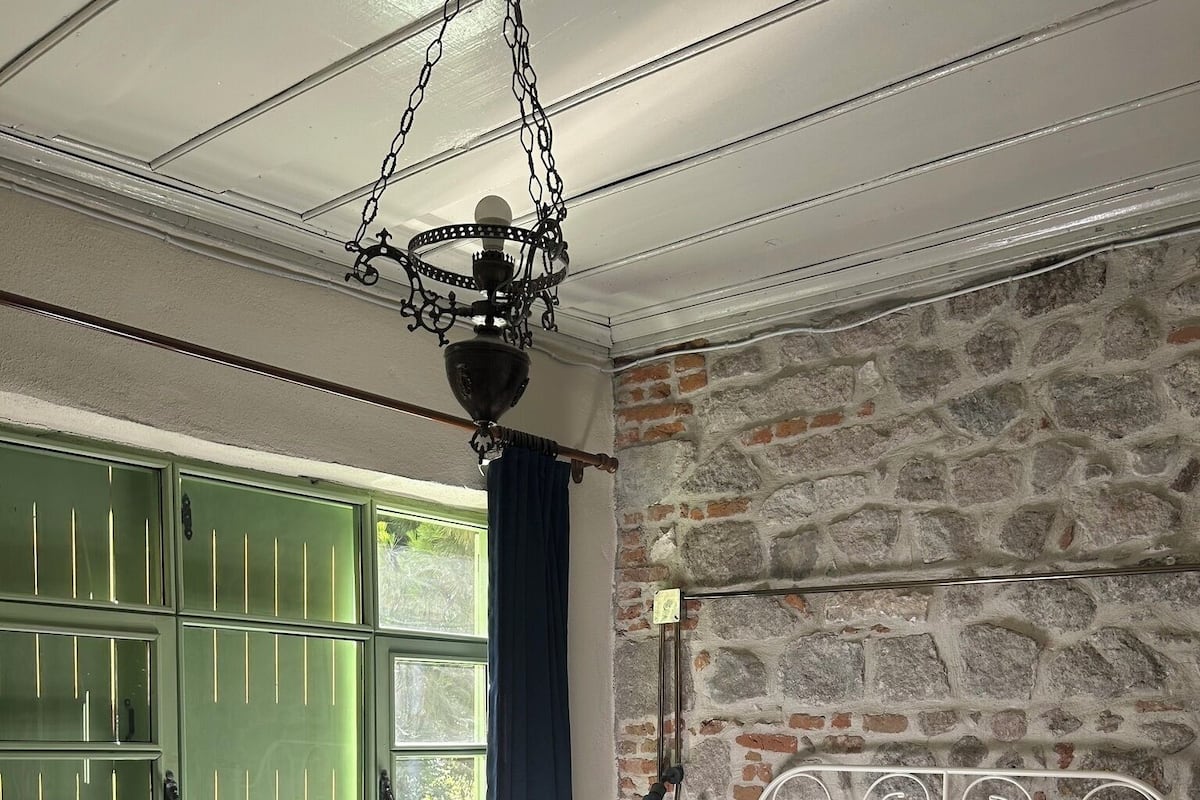 Un dormitorio con pared de piedra, estructura de cama de metal, ventana verde y lámpara colgante.