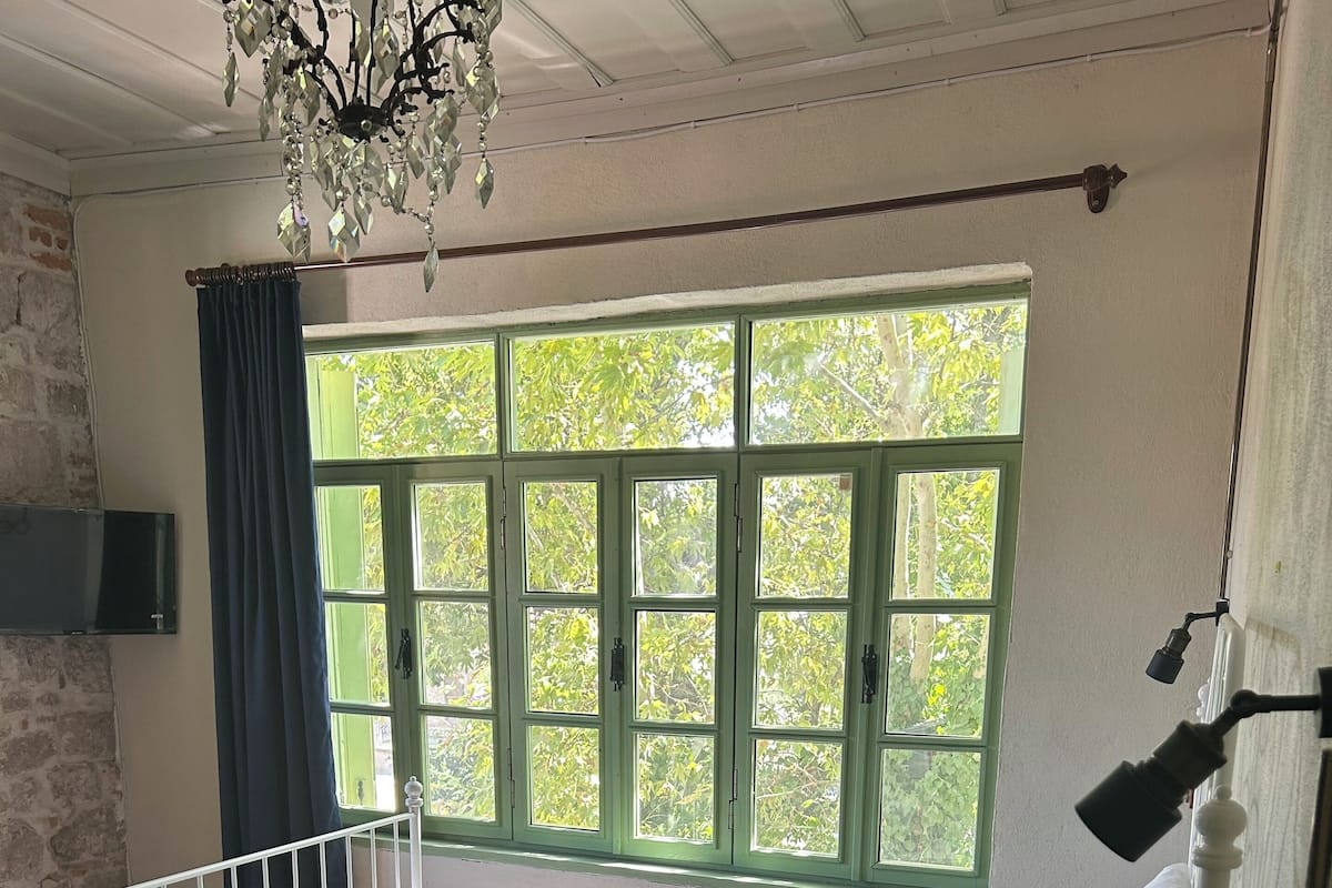Una habitación con una cama, un ventanal con contraventanas verdes, una araña de cristal y una puerta con manija.