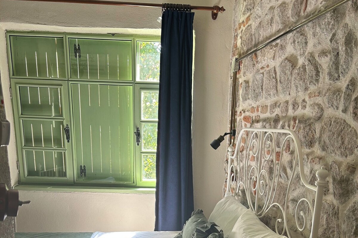 Een bed met witte beddengoed en decoratieve kussens, een groen raam met tralies, een blauw gordijn, en een stenen muur.