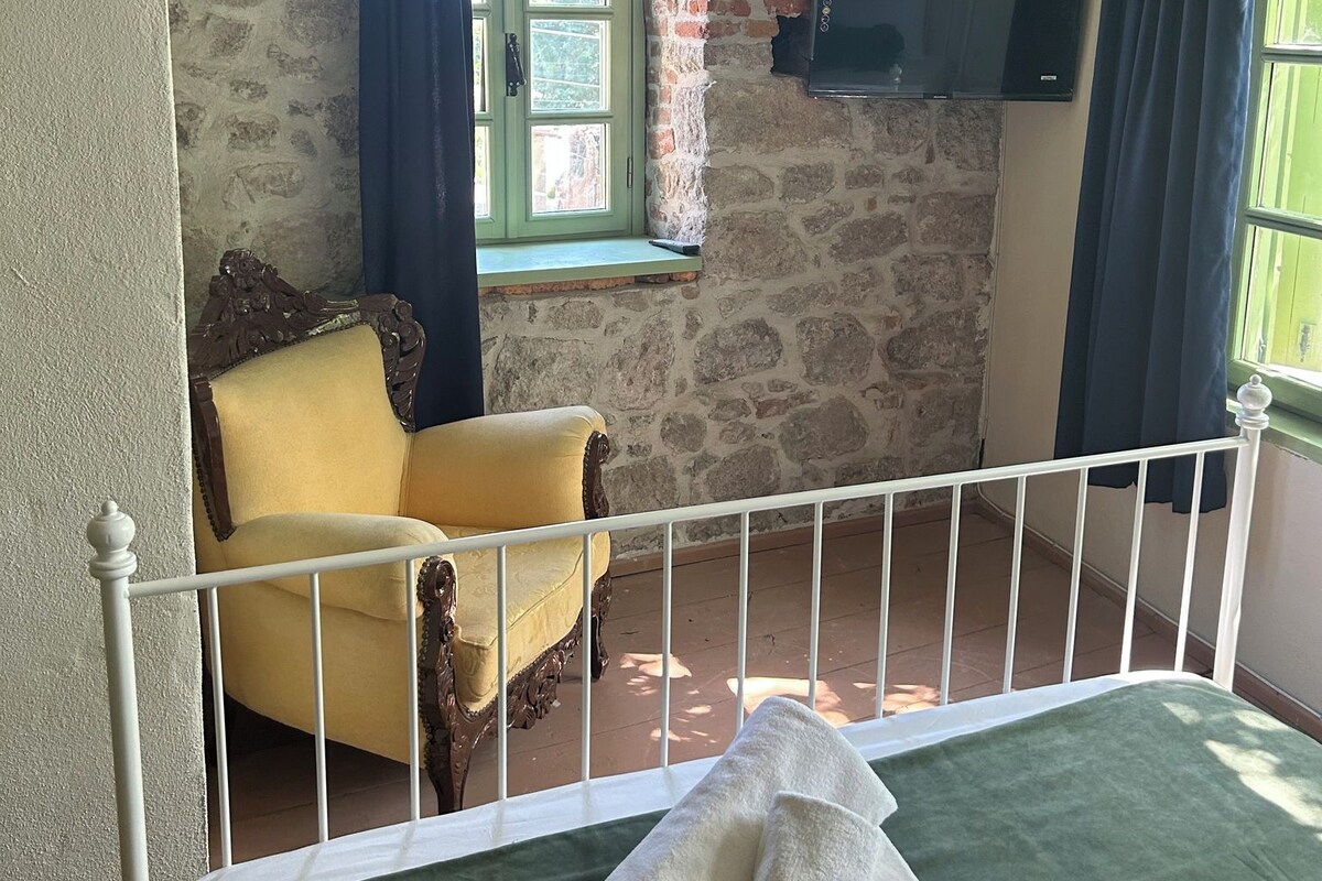 Een kamer met een stenen muur, een raam met gordijnen, een gele fauteuil, een bed met een groene sprei en een witte metalen balustrade.