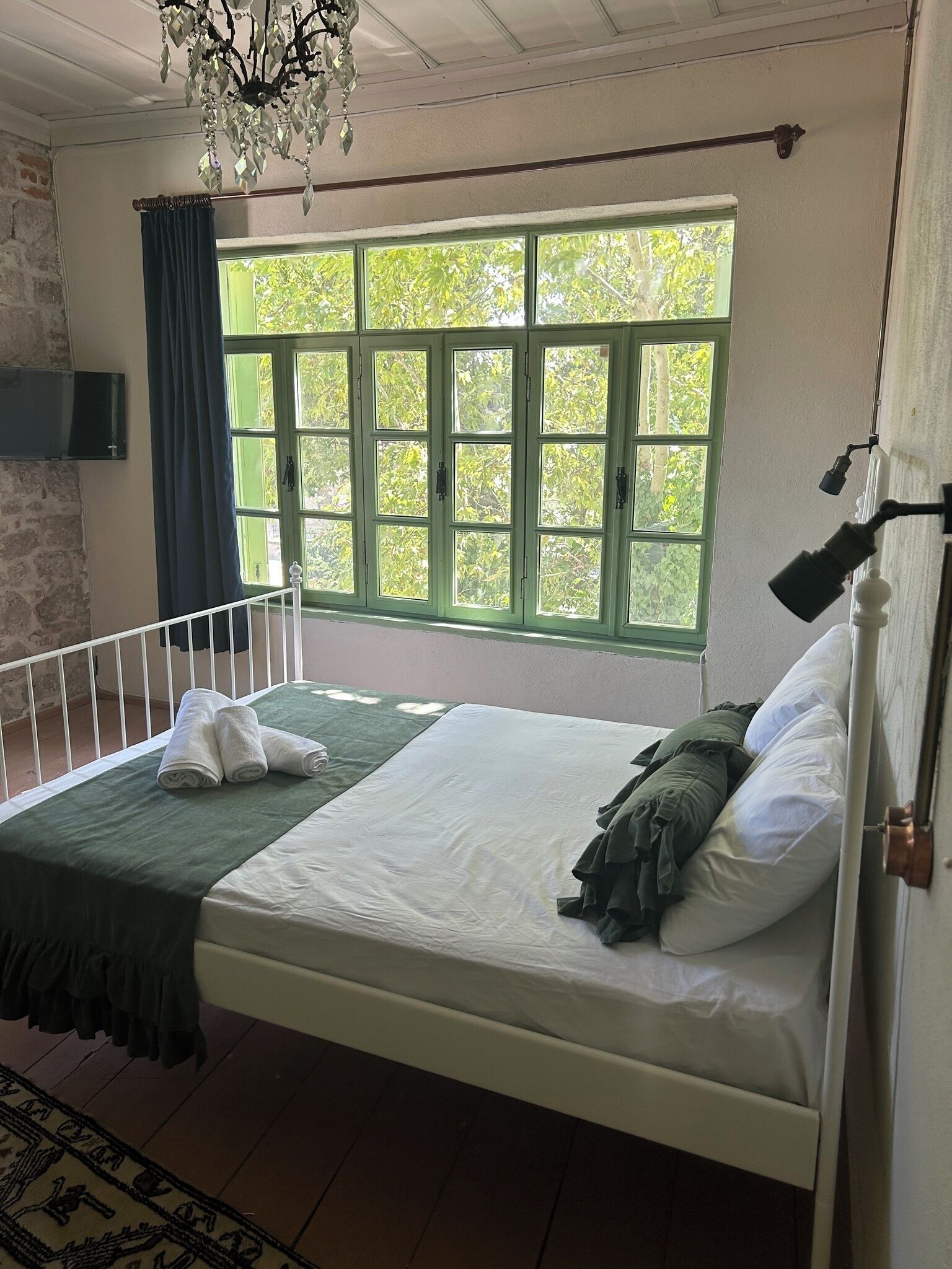 Een kamer met een bed, een raam met groene luiken, een kroonluchter en een aan de muur bevestigde televisie.