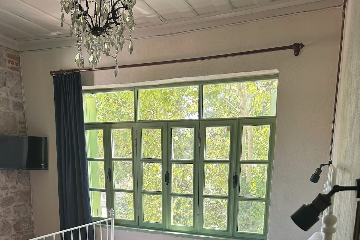 Una habitación con cama, ventana con contraventanas verdes, una lámpara de araña y televisor montado en la pared.