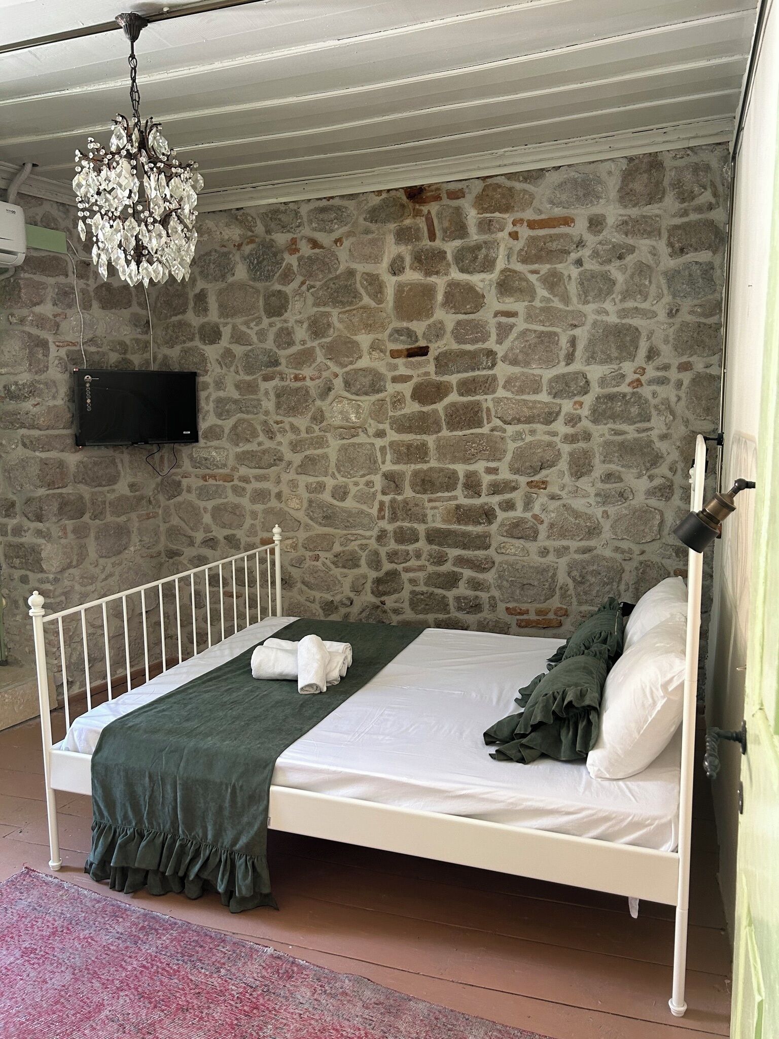 Een bed met een wit frame, een groene spreideken en een donkergroene plaid. Een kroonluchter hangt aan het plafond, en een flatscreen televisie is bevestigd aan de stenen muur.