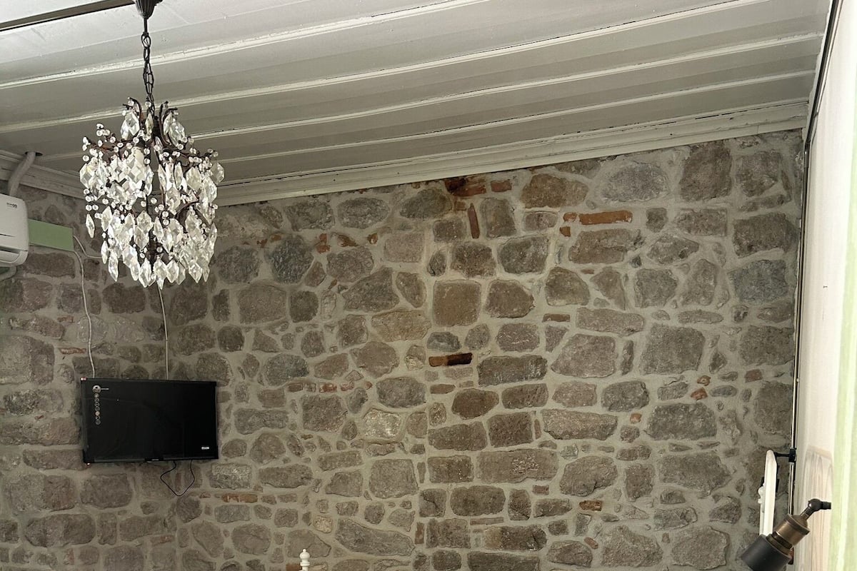 Una cama con estructura blanca, cubrecama verde y manta verde oscuro. Un candelabro cuelga del techo, y un televisor de pantalla plana está montado en la pared de piedra.