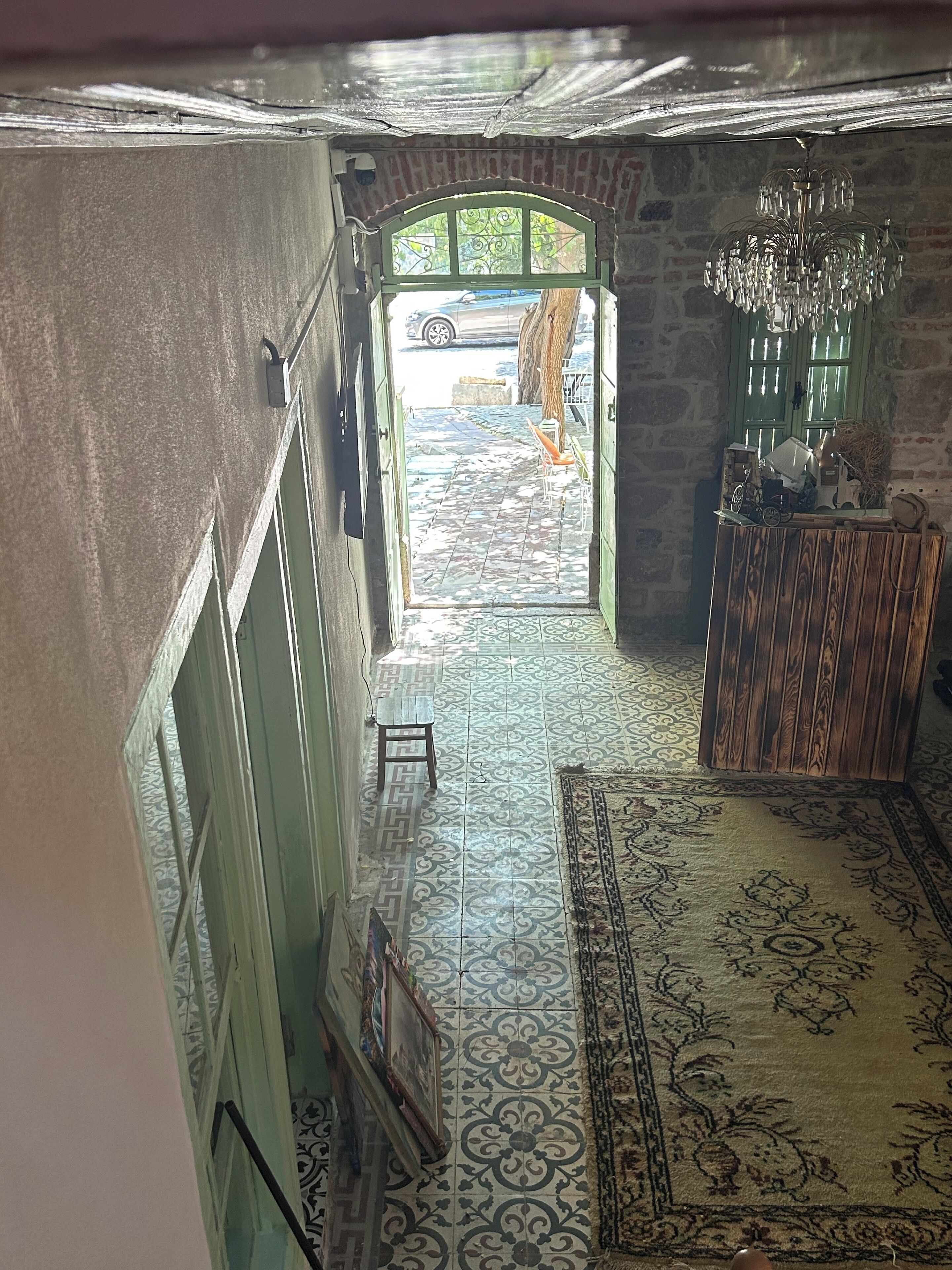 A hallway with patterned floor tiles, a wooden bench, a chandelier, and a view through an open doorway to an outdoor area with a tree and a parked car.
