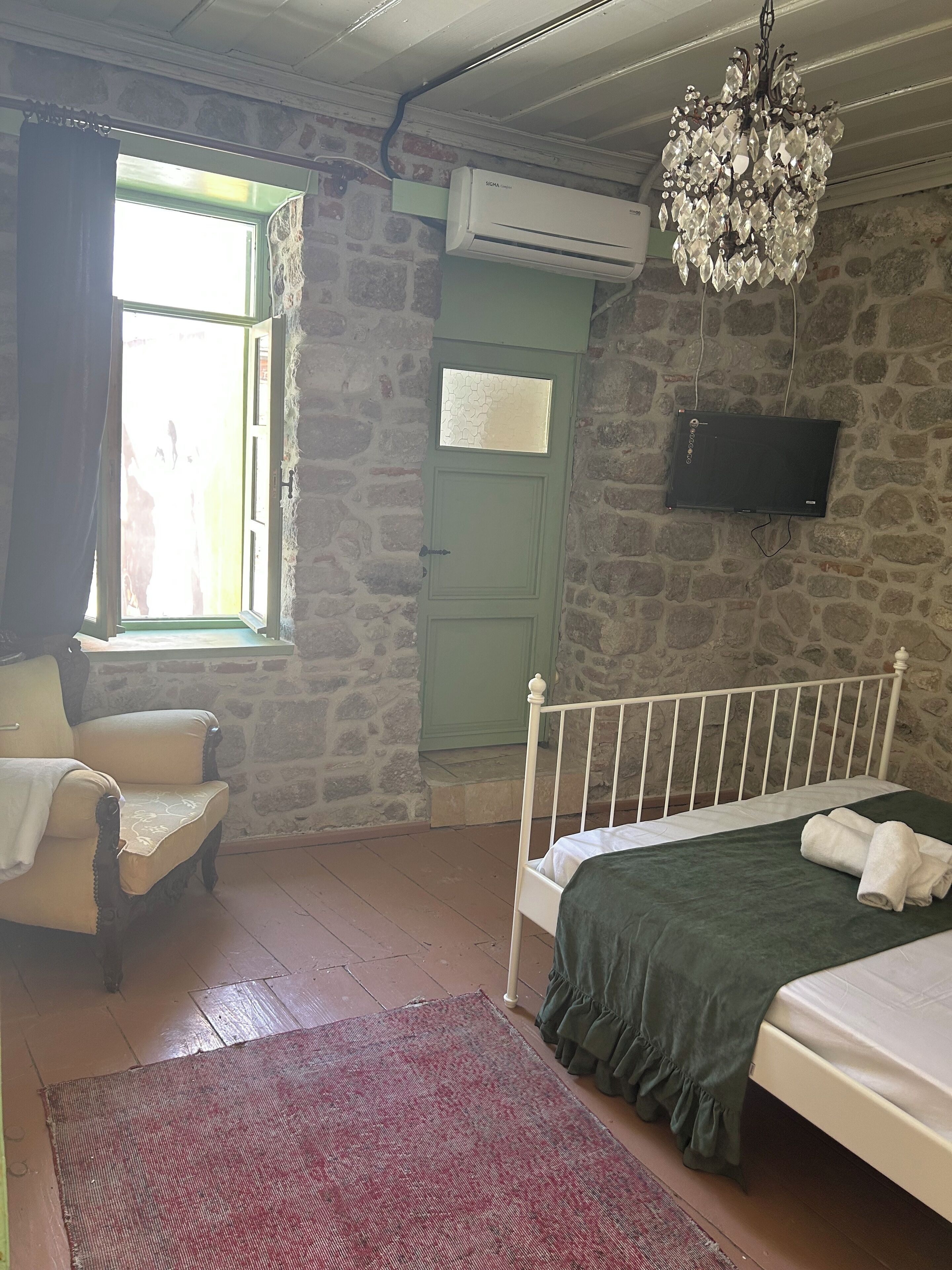 A bedroom with a stone wall, a green door, a white bed, a chair, a red rug, and a chandelier.