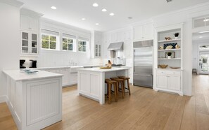 A modern kitchen with white cabinetry, a large island with stools, a stainless steel refrigerator, and a window overlooking greenery.
