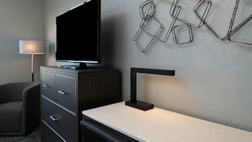 Une chambre d’hôtel moderne équipée d’un téléviseur à écran plat posé sur un meuble, d’un bureau avec une lampe, d’une chaise et d’une décoration murale aux motifs géométriques.