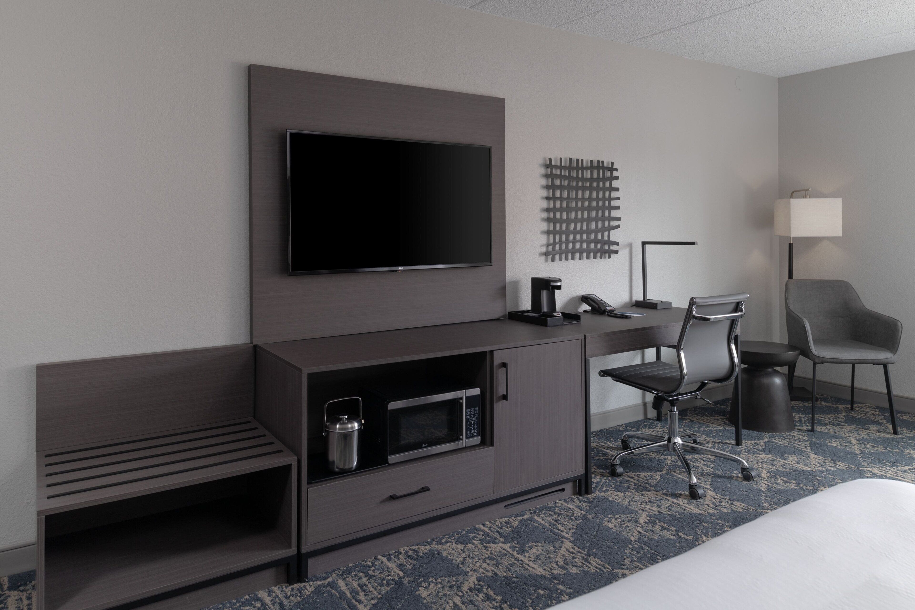 Una habitación de hotel moderna con una televisión de pantalla plana montada en la pared, un escritorio con una silla, un microondas y un mueble auxiliar.