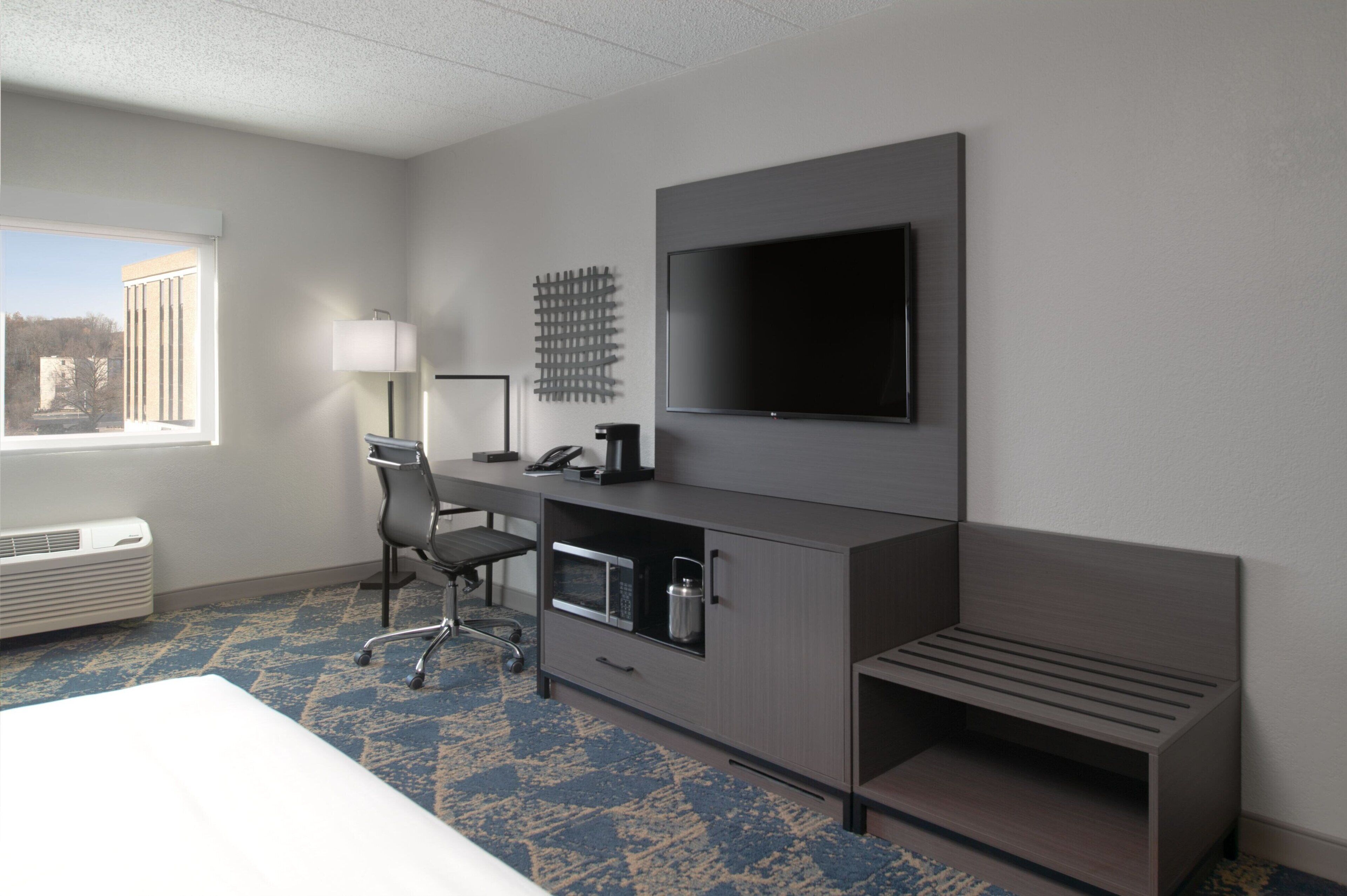 Habitación de hotel con una cama grande, un escritorio con silla, televisor de pantalla plana montado en la pared, microondas, cafetera y ventana con vistas a edificios.