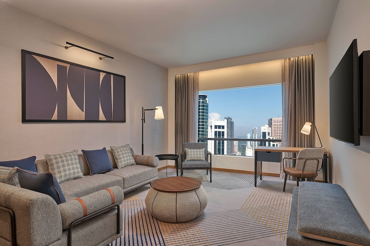 Een moderne hotelkamer met een bank, fauteuil, rond bijzettafeltje en een bureau met stoel. Een groot raam biedt uitzicht over de stad.