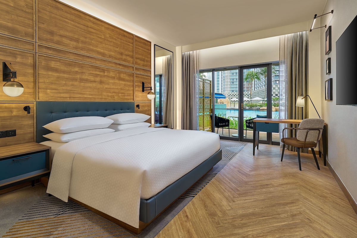 Een moderne hotelkamer met een groot bed, een bureau en uitzicht op het zwembad en de omliggende gebouwen.